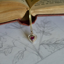Heart-shaped pendant with a red gemstone on a white surface with engraved designs, next to an open book.