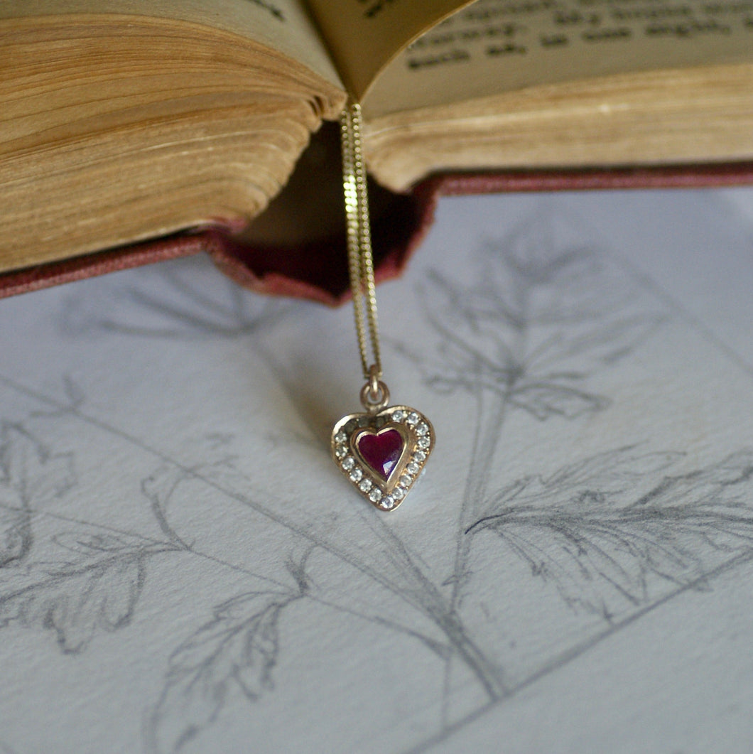 Heart-shaped pendant with a red gemstone on a white surface with engraved designs, next to an open book.