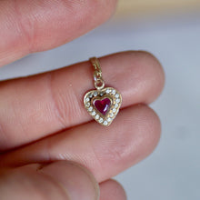 Heart-shaped pendant with a red gemstone held between fingers against a blurred background